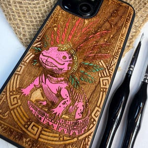 May include: A wooden phone case with a laser-engraved design of a pink axolotl wearing a feathered headdress. The axolotl is surrounded by detailed patterns. The case has a black border. Two black tools are next to the case.