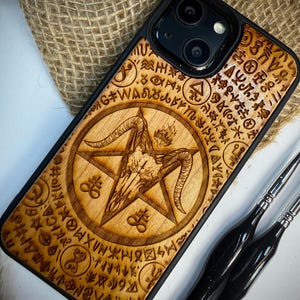 May include: A wooden phone case with a black border. The case features a detailed engraving of a pentagram with a goat skull at the center, surrounded by symbols. The wood has a light brown color.