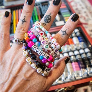 May include: A hand adorned with multiple beaded bracelets in various colors, spelling out "QUEER" and other words. The bracelets are stacked on the wrist and fingers, alongside a silver ring. The background includes a red art supply case.