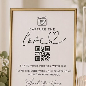 May include: A gold-framed sign with white background featuring a black camera icon, the text CAPTURE THE love, a QR code, the text SHARE YOUR PHOTOS WITH US! SCAN THE CODE WITH YOUR SMARTPHONE & UPLOAD YOUR PHOTOS, and the names Sarah & Jonas.