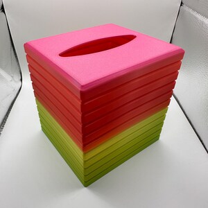 May include: A square tissue box with a gradient color scheme. The top is a bright pink, transitioning to red, and then to a yellow-green at the base. The top has an oval opening for tissue dispensing.