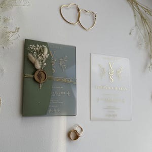 May include: A wedding invitation suite with a sage green invitation with gold detailing, a clear invitation, gold heart-shaped hoop earrings, and a gold ring. The invitation is decorated with dried flowers and a wax seal.