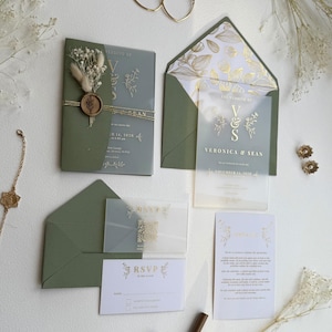 May include: An olive green wedding invitation suite with gold foil accents. The suite includes invitations, RSVP cards, and envelopes. The main invitation is adorned with a dried flower arrangement and a wax seal. Gold hoop earrings and floral accents complete the presentation.