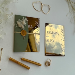 Può includere: Una collezione di articoli di cancelleria e accessori per matrimoni. Include orecchini a cerchio dorati a forma di cuore, due penne dorate, un anello dorato e due inviti a specchio con nomi e date. Un invito è verde oliva.