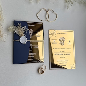 Può includere: Un set di inviti di nozze con uno schema di colori blu navy e oro. Il set include un invito blu navy con un accento dorato, un biglietto d'oro per salvare la data, orecchini a cerchio dorati a forma di cuore e fedi nuziali.