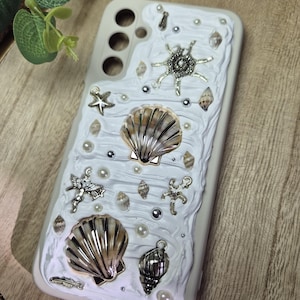 Może przedstawiać: Białe etui na telefon zdobione muszlami, rozgwiazdami, perłami i srebrnymi zawieszkami. Etui ma teksturowany, falisty wzór z różnymi ozdobami z muszli i srebrnymi akcentami.