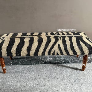 May include: A rectangular bench featuring a textured, woven fabric top with a black and beige zebra stripe pattern. The bench is supported by four turned, dark brown wooden legs. The text juterugscarpets is visible above the bench.