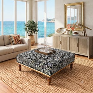 Puede incluir: Un otomán cuadrado con una tela oscura que presenta un denso patrón de flores y enredaderas claras, sostenido por cuatro patas de madera torneadas. Una bandeja rectangular de madera con cristalería transparente descansa encima.
