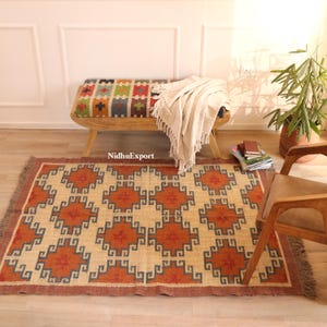 May include: A rectangular beige rug featuring a geometric design in orange, brown, and blue. The rug has a fringed edge and is placed on a wooden floor. A wooden bench with a colourful patterned seat and a white blanket are in the background.
