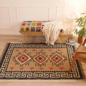 Op de afbeelding: Een rechthoekig beige tapijt met een zwarte rand en geometrische patronen. Het tapijt heeft een herhalend ontwerp van ruiten en driehoeken in rood, zwart en beige. Op de achtergrond een houten bank met een kleurrijk patroon en een crèmekleurige deken.