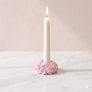 May include: A lit white taper candle in a pink floral candle holder. The candle holder is decorated with pink and light blue flowers. The background is a soft pink colour, and the candle is centered in the frame.