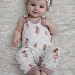 Könnte beinhalten: Ein Baby in einem weißen Strampler und passendem Stirnband, beide mit einem Ballerina-Print in Rosa und Braun. Der Strampler hat rosa Verzierungen und Träger. Das Baby liegt auf einer weichen, weißen Decke.