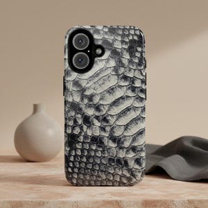 Pode incluir: Capa de smartphone com um padrão texturizado de pele de cobra em tons de cinza e branco. A capa tem um recorte para uma câmera de lente dupla e botões laterais pretos. O fundo inclui um pequeno vaso e um pano cinza dobrado.
