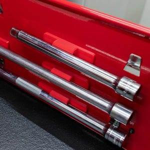 May include: A red toolbox interior with three chrome extension bars and sockets. The tools are held in place by red plastic dividers. The background is a dark gray surface.