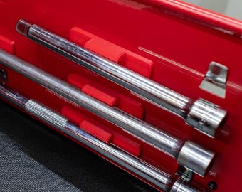 May include: A red toolbox interior with three chrome extension bars and sockets. The tools are held in place by red plastic dividers. The background is a dark grey surface.