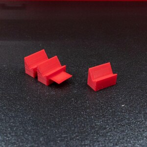 May include: Several small, bright red, 3D-printed objects are arranged on a dark, textured surface. The objects appear to be small, angled stands or supports, possibly for holding items. The lighting highlights their geometric shapes.