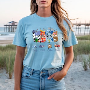 Puede incluir: Una camiseta de manga corta azul claro con un estampado gr&aacute;fico de la palabra mama en letras grandes y coloridas decoradas con varios personajes de coches de dibujos animados, banderas a cuadros y estrellas, con cuatro nombres en cursiva.