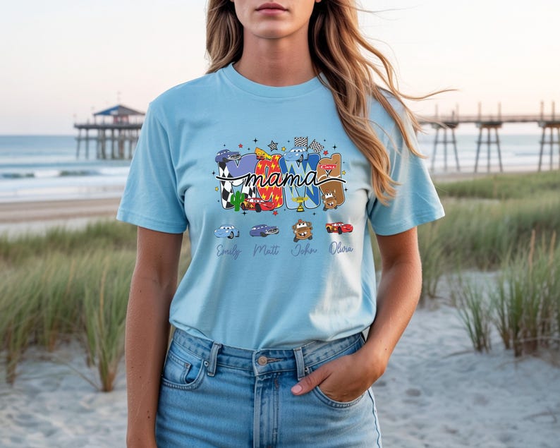 Puede incluir: Una camiseta de manga corta azul claro con un estampado gr&aacute;fico de la palabra mama en letras grandes y coloridas decoradas con varios personajes de coches de dibujos animados, banderas a cuadros y estrellas, con cuatro nombres en cursiva.