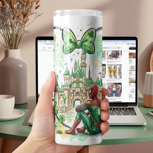 May include: A white tumbler featuring a green bow with shamrocks, a castle, and a seated Spider-Man in a green and red suit. The design includes fireworks and a green and white color scheme.