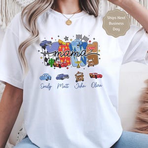 Puede incluir: Una camiseta blanca de manga corta con un gr&aacute;fico colorido que muestra la palabra mama en letra cursiva. Las letras est&aacute;n rellenas con varios personajes de coches de dibujos animados, banderas a cuadros y estrellas. Debajo del gr&aacute;fico hay cuatro iconos de coches peque&ntilde;os con los nombres Emily, Matt, John y Olivia impresos en texto azul cursivo.