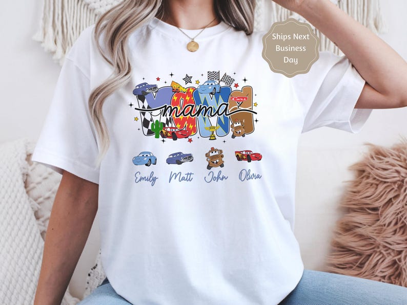 Puede incluir: Una camiseta blanca de manga corta con un gr&aacute;fico colorido que muestra la palabra mama en letra cursiva. Las letras est&aacute;n rellenas con varios personajes de coches de dibujos animados, banderas a cuadros y estrellas. Debajo del gr&aacute;fico hay cuatro iconos de coches peque&ntilde;os con los nombres Emily, Matt, John y Olivia impresos en texto azul cursivo.