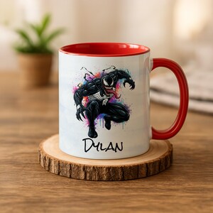 Op de afbeelding: Witte keramische mok met een rode binnenkant en oor. De mok heeft een kleurrijke afbeelding van het Marvel Comics-personage Venom, met de naam "Dylan" eronder. De mok staat op een houten onderzetter.