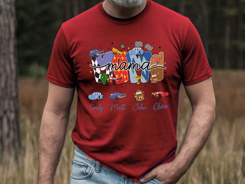 Puede incluir: Una camiseta roja de manga corta con un dise&ntilde;o gr&aacute;fico que muestra la palabra mama en cursiva sobre letras grandes que forman MOM. El dise&ntilde;o incluye varios personajes de coches de dibujos animados, banderas a cuadros y los nombres Emily, Matt, John y Olura.
