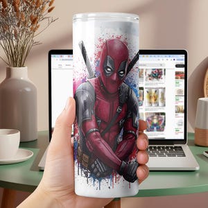 K&ouml;nnte beinhalten: Ein wei&szlig;er Becher mit einer lebendigen Illustration von Deadpool in seinem roten und schwarzen Anzug, komplett mit Schwertern. Das Design enth&auml;lt blaue und violette Farbspritzer, die eine dynamische &Auml;sthetik erzeugen. Ideal f&uuml;r Fans der Marvel-Figur.