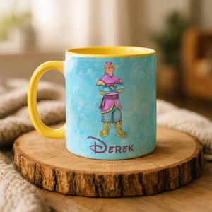 Peut inclure: Une tasse en c&eacute;ramique avec une anse et un int&eacute;rieur jaunes. L'ext&eacute;rieur pr&eacute;sente un motif de style aquarelle avec un personnage de dessin anim&eacute; en tenue violette et bleue, avec le nom "Derek" en &eacute;criture violette. La tasse est pos&eacute;e sur une surface en bois.