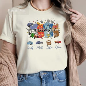Puede incluir: Una camiseta de color crema con un estampado gr&aacute;fico que incluye la palabra mama en letra cursiva. El dise&ntilde;o presenta coloridos coches de carreras de dibujos animados, banderas a cuadros y los nombres Emily, Matt, John y Olivia.