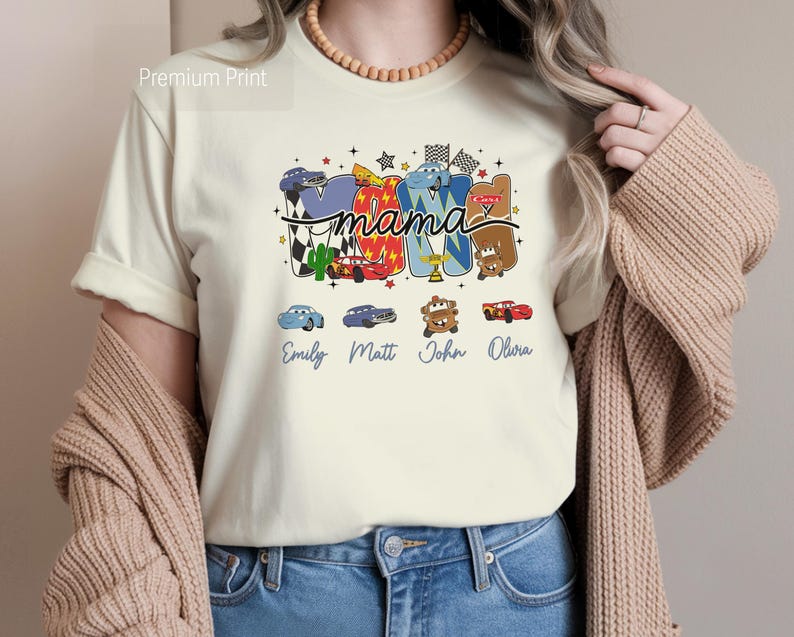 Puede incluir: Una camiseta de color crema con un estampado gr&aacute;fico que incluye la palabra mama en letra cursiva. El dise&ntilde;o presenta coloridos coches de carreras de dibujos animados, banderas a cuadros y los nombres Emily, Matt, John y Olivia.