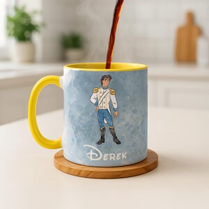 May include: A ceramic mug with a yellow handle and interior, featuring a cartoon illustration of a prince in a white and blue uniform. The mug has the name "DEREK" printed on it and sits on a wooden coaster. Coffee is being poured into the mug.