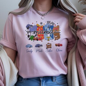 Puede incluir: Una camiseta rosa claro con un dise&ntilde;o gr&aacute;fico que incluye la palabra mama en letra cursiva. Las letras est&aacute;n rellenas con varios personajes de coches de dibujos animados, banderas a cuadros y estrellas. Debajo hay cuatro nombres: Emily, Matt, John y Olivia.