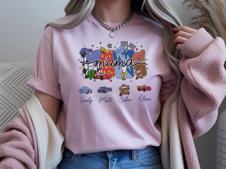 Puede incluir: Una camiseta rosa claro con un dise&ntilde;o gr&aacute;fico que incluye la palabra mama en letra cursiva. Las letras est&aacute;n rellenas con varios personajes de coches de dibujos animados, banderas a cuadros y estrellas. Debajo hay cuatro nombres: Emily, Matt, John y Olivia.