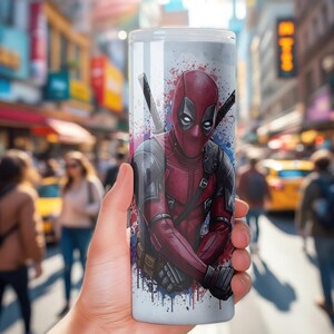K&ouml;nnte beinhalten: Ein wei&szlig;er Becher mit einer lebendigen Illustration von Deadpool in seinem charakteristischen rot-schwarzen Anzug, mit zwei Katanas auf dem R&uuml;cken. Das Design umfasst Farbspritzer in Blau und Lila. Der Becher wird vor einem verschwommenen Stadtbild gehalten.