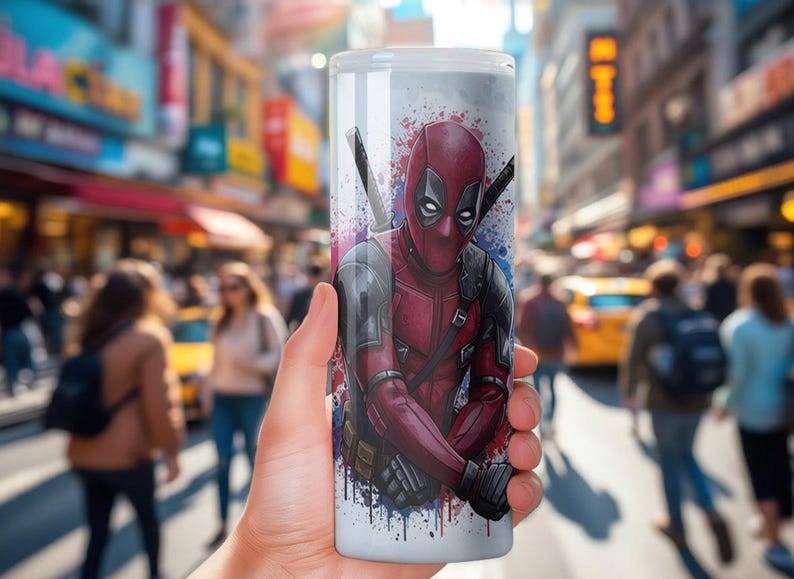 K&ouml;nnte beinhalten: Ein wei&szlig;er Becher mit einer lebendigen Illustration von Deadpool in seinem charakteristischen rot-schwarzen Anzug, mit zwei Katanas auf dem R&uuml;cken. Das Design umfasst Farbspritzer in Blau und Lila. Der Becher wird vor einem verschwommenen Stadtbild gehalten.