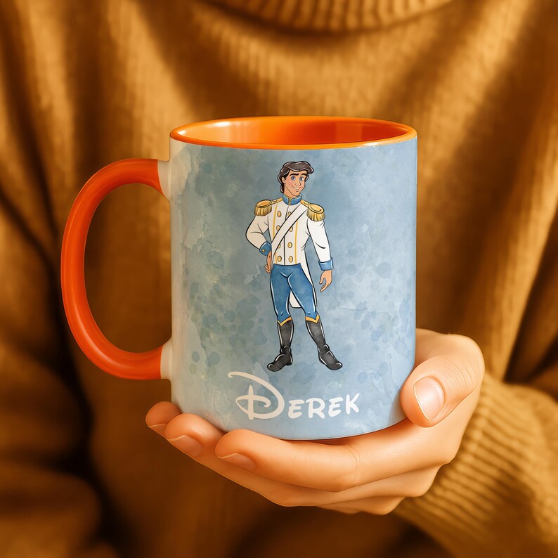May include: A ceramic mug with an orange handle and interior. The exterior features a watercolor-style design with a cartoon illustration of a prince in a white and blue uniform. The name "Derek" is printed on the mug.