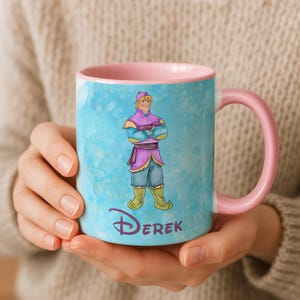 Peut inclure: Une tasse en c&eacute;ramique avec un int&eacute;rieur et une anse roses, orn&eacute;e d'une illustration &agrave; l'aquarelle d'un personnage v&ecirc;tu d'une tenue violette et bleue. Le nom du personnage, "Derek", est imprim&eacute; en violet en dessous. La tasse a un fond bleu clair.