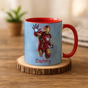 Może przedstawiać: Ceramiczny kubek z czerwonym wnętrzem i uchwytem, z ilustracją Iron Mana na jasnoniebieskim tle. Projekt zawiera imię "Danny" w białym tekście. Kubek stoi na drewnianej podstawce.
