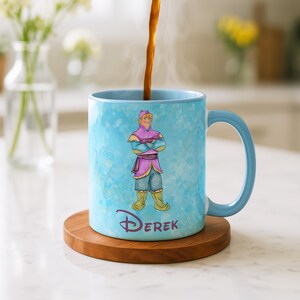 Peut inclure: Une tasse en c&eacute;ramique bleu clair avec une illustration &agrave; l'aquarelle d'un personnage et le nom "Derek" en police Disney. La tasse a une anse et un bord bleu clair, et repose sur un dessous de verre en bois. Du caf&eacute; est vers&eacute; dans la tasse.