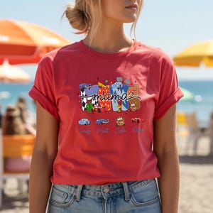 Puede incluir: Una camiseta de color coral con un dise&ntilde;o gr&aacute;fico que presenta la palabra mama en letras grandes y estilizadas rellenas con varios patrones y personajes de tem&aacute;tica automovil&iacute;stica. Debajo de la palabra, cuatro coches peque&ntilde;os est&aacute;n impresos sobre los nombres Emily, Matt, John y Olive.