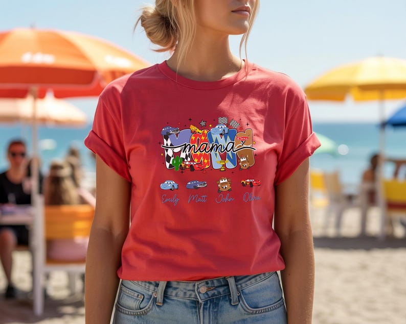 Puede incluir: Una camiseta de color coral con un dise&ntilde;o gr&aacute;fico que presenta la palabra mama en letras grandes y estilizadas rellenas con varios patrones y personajes de tem&aacute;tica automovil&iacute;stica. Debajo de la palabra, cuatro coches peque&ntilde;os est&aacute;n impresos sobre los nombres Emily, Matt, John y Olive.