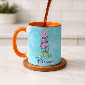 Peut inclure: Mug en c&eacute;ramique avec une anse et un int&eacute;rieur orange, orn&eacute; d'une illustration de personnage de dessin anim&eacute; sur fond bleu clair. Le personnage porte des v&ecirc;tements violets et bleus. Le nom "DEREK" est imprim&eacute; en bas.
