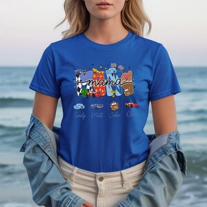 Puede incluir: Una camiseta de manga corta color azul real con un estampado gr&aacute;fico de la palabra MAMA en letras de bloque coloridas y estampadas. El dise&ntilde;o incluye personajes de coches de dibujos animados, banderas a cuadros y los nombres debajo.