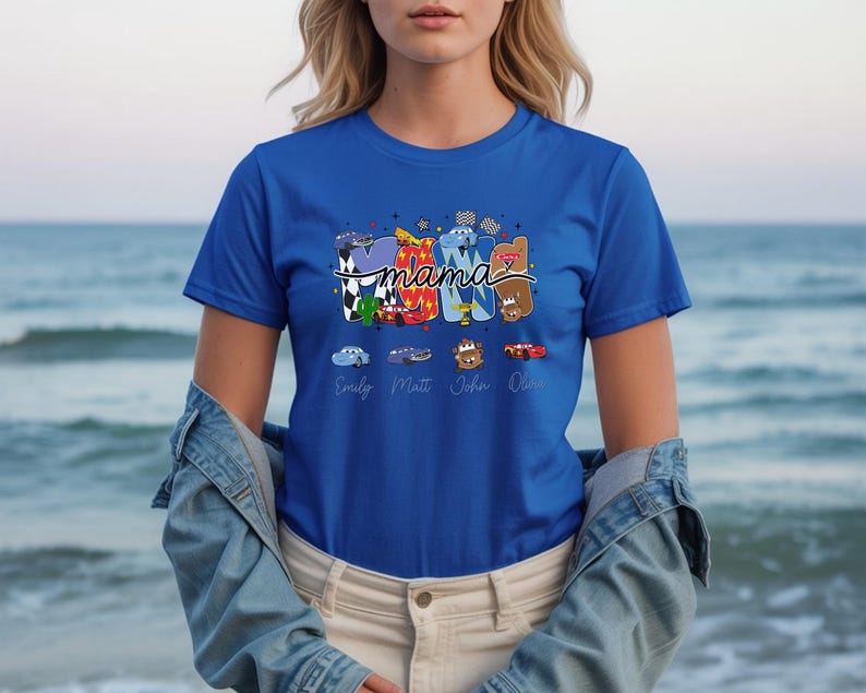 Puede incluir: Una camiseta de manga corta color azul real con un estampado gr&aacute;fico de la palabra MAMA en letras de bloque coloridas y estampadas. El dise&ntilde;o incluye personajes de coches de dibujos animados, banderas a cuadros y los nombres debajo.