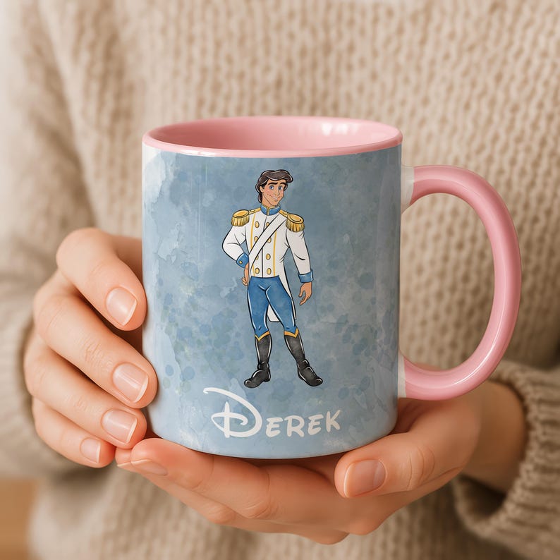 May include: A ceramic mug with a pink handle and interior. The exterior features a watercolor-style blue background with a cartoon illustration of a prince in a white and blue uniform. The name "Derek" is printed in white script below the illustration.