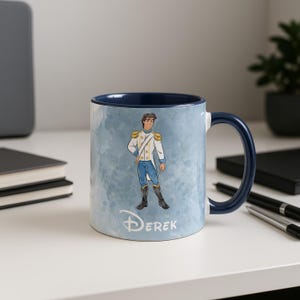 May include: A ceramic mug with a blue interior and handle. The exterior features a watercolor-style background and a cartoon illustration of a prince in a white and blue uniform. The name "Derek" is printed in a script font.