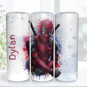 K&ouml;nnte beinhalten: Ein wei&szlig;er Becher mit einem Deadpool-Design. Die Figur tr&auml;gt ein rot-schwarzes Kost&uuml;m vor einem farbenfrohen Aquarell-Spritzer-Hintergrund. Der Name "Dylan" steht in Rot geschrieben.