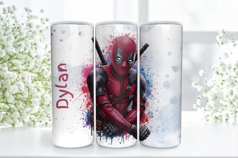K&ouml;nnte beinhalten: Ein wei&szlig;er Becher mit einem Deadpool-Design. Die Figur tr&auml;gt ein rot-schwarzes Kost&uuml;m vor einem farbenfrohen Aquarell-Spritzer-Hintergrund. Der Name "Dylan" steht in Rot geschrieben.