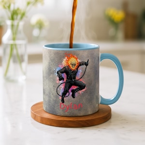 以下が含まれることがあります： 内側とハンドルが水色のセラミックコーヒーマグ。炎のドクロのキャラクターのグラフィックが特徴です。マグカップは木製のコースターの上にあり、前面には「Dylan」という名前が印刷されています。コーヒーがマグカップに注がれています。